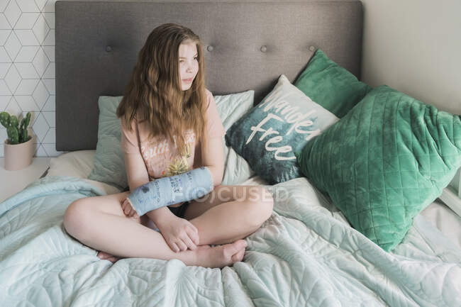 Junges Mädchen sitzt mit dem Arm in einem blauen Gips auf einem Bett — Stockfoto
