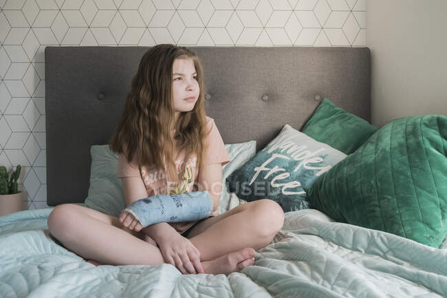 Jeune fille assise jambes croisées sur un lit avec son bras dans un plâtre — Photo de stock