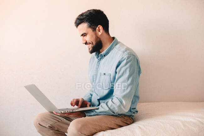 Hombre feliz usando el ordenador portátil mientras está sentado en casa contra la pared - foto de stock