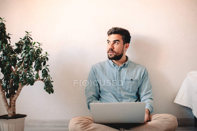 Hombre usando el ordenador portátil mientras está sentado contra la pared en el suelo - foto de stock