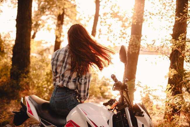 Mujer joven sacudiendo el pelo mientras se relaja sentado en motocicleta por el río - foto de stock
