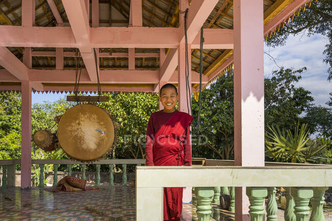 Улыбающийся буддийский монах, стоящий рядом с гонгом на террасе монастыря в провинции Луанг Намтха, Лаос — стоковое фото