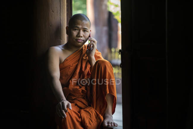 Буддийский монах, одетый в оранжевый халат, звонит по мобильному телефону, сидя в окне, Мандалай, Мьянма — стоковое фото