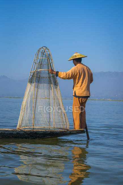 Vue arrière du pêcheur d'Intha debout avec un filet de pêche conique typique sur le bateau contre un ciel bleu clair, lac Inle, Nyaungshwe, Myanmar — Photo de stock