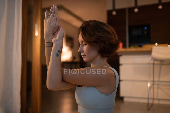 Side view of young female meditating with Eagle arms and closed eyes at night at home — Stock Photo