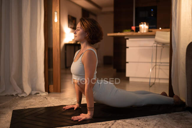 Side view of fit female doing Upward Facing Dog pose with closed eyes at night at home — Stock Photo