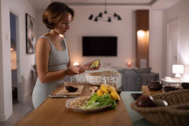 Тонкая женщина добавляет спелый авокадо в салат во время приготовления здорового ужина на кухне — стоковое фото