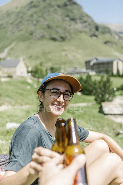 Femme voyageuse trinquant à la bière avec un homme anonyme assis au bord d'un lac dans les montagnes — Photo de stock