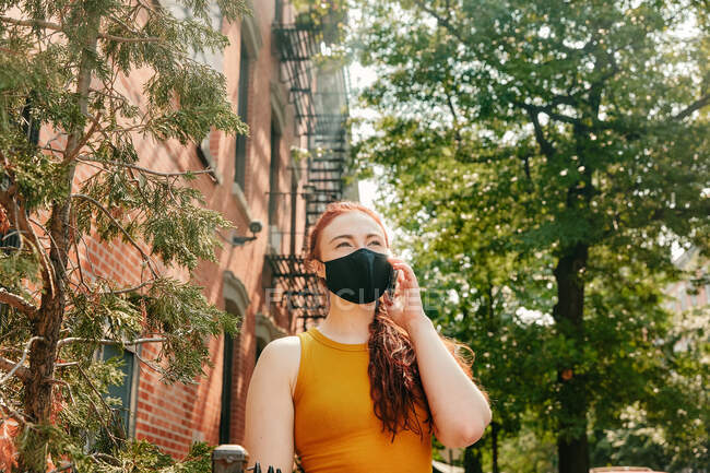 Mujer joven en el teléfono con mascarilla en la calle Brooklyn por los árboles - foto de stock