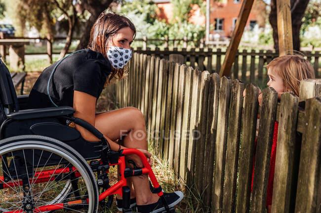 Madre en silla de ruedas con una máscara médica mirando a su pequeño papá - foto de stock