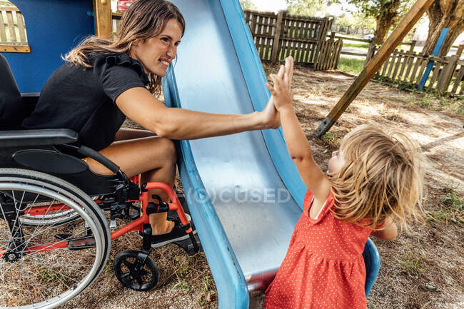 Madre en silla de ruedas celebrando con su hija pequeña en un parque - foto de stock