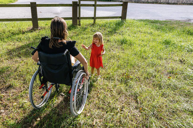 Маленька дівчинка грає на зеленому полі з матір'ю на інвалідному візку — стокове фото