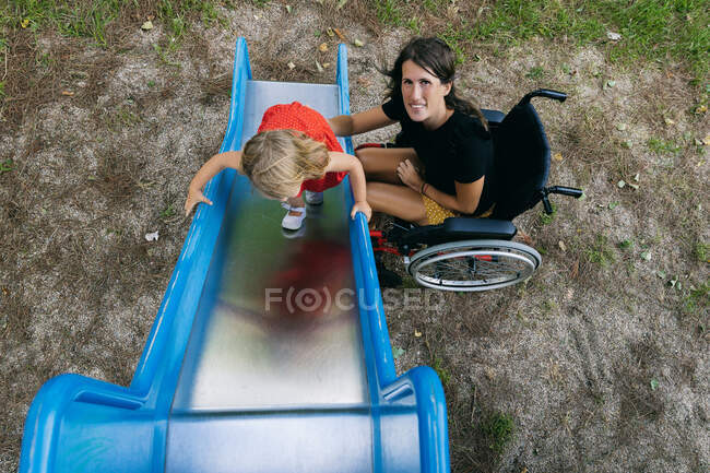 Sorrindo mãe em uma cadeira de rodas brincando com sua filhinha em um s — Fotografia de Stock