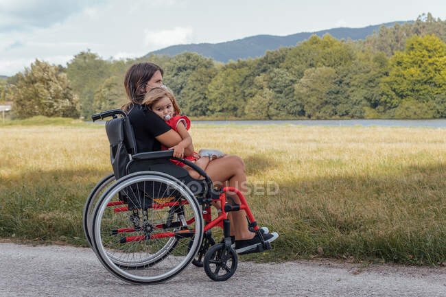 Mãe em uma cadeira de rodas abraçando sua filhinha com um humor de chupeta — Fotografia de Stock