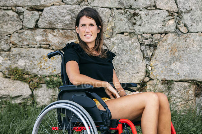 Mujer joven sonriendo en una silla de ruedas con una pared de piedras en la espalda - foto de stock