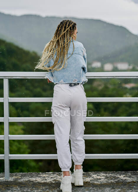 Face arrière d'une jeune femme aux cheveux blonds tressés portant une veste en denim et un jean blanc reposant sur un barrage — Photo de stock