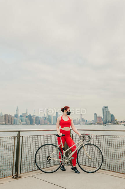 Молодая велосипедистка в маске для лица с велосипедом на набережной — стоковое фото