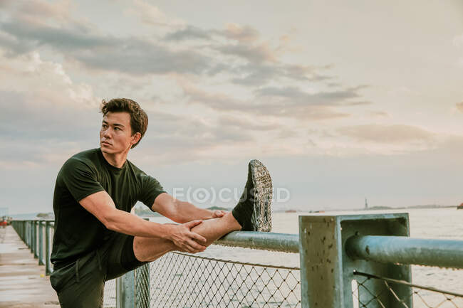 Atleta masculino que se estende à beira-mar durante o pôr do sol — Fotografia de Stock