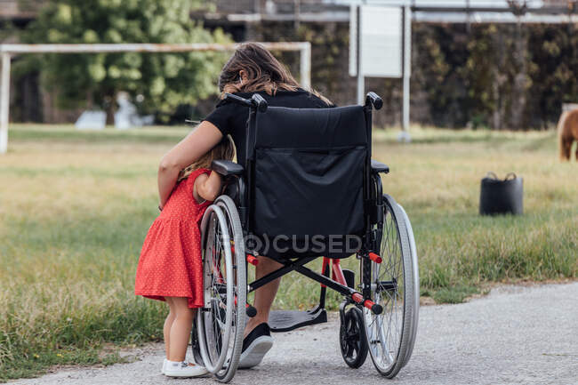 Madre en silla de ruedas abrazando a su hijita con un campo verde - foto de stock