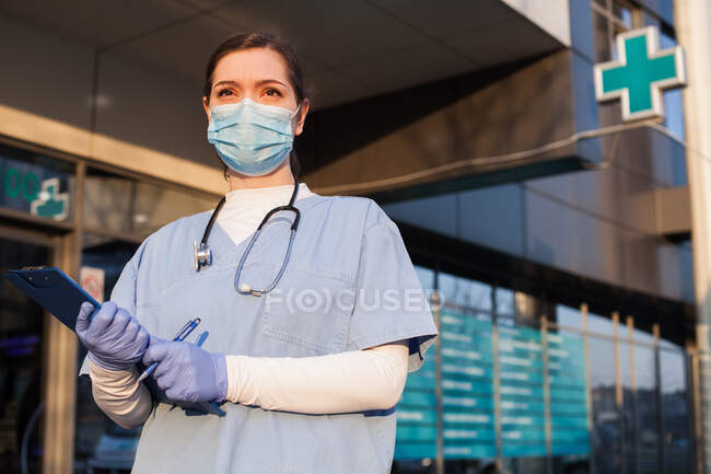 Jovem médica em pé em frente à unidade de saúde, usando máscara protetora e equipamento de EPI, segurando prancheta de paciente médico, crise pandêmica COVID-19 — Fotografia de Stock