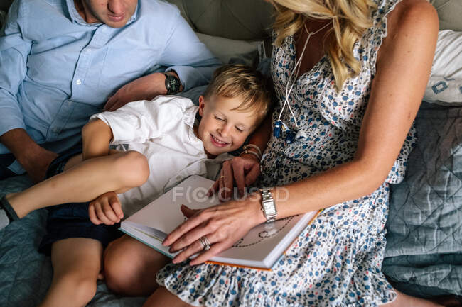 Happy young boy snuggling his parents while they read on bed — Stock Photo