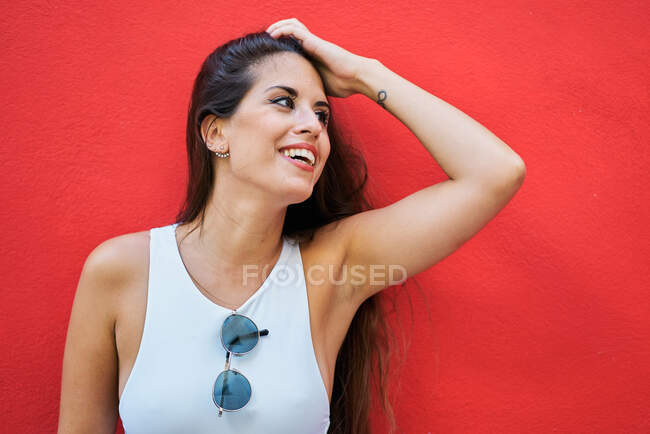 Jeune femme aux cheveux longs portant des lunettes de soleil élégantes et debout près du mur de bâtiment rouge en ville — Photo de stock