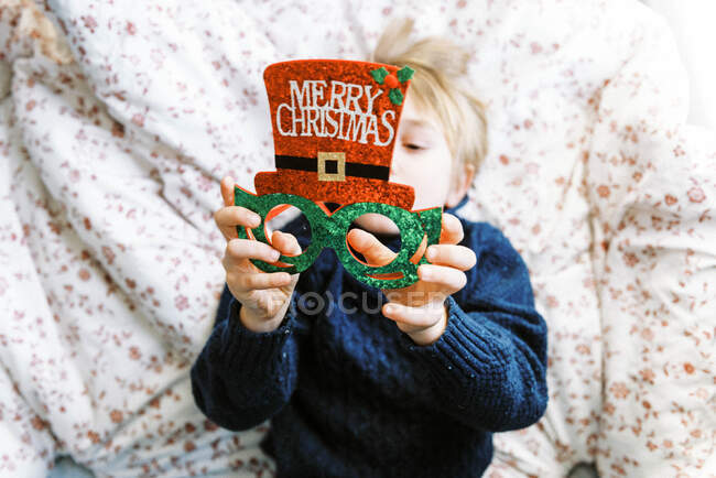 Little boy in a knit sweater looking at Christmas glasses lying on bed — Stock Photo