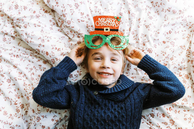 Petit garçon dans un pull en tricot avec des lunettes de Noël couché sur le lit — Photo de stock
