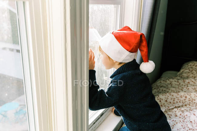 Menino com chapéu de Natal a olhar pela janela para a cláusula do Pai Natal — Fotografia de Stock