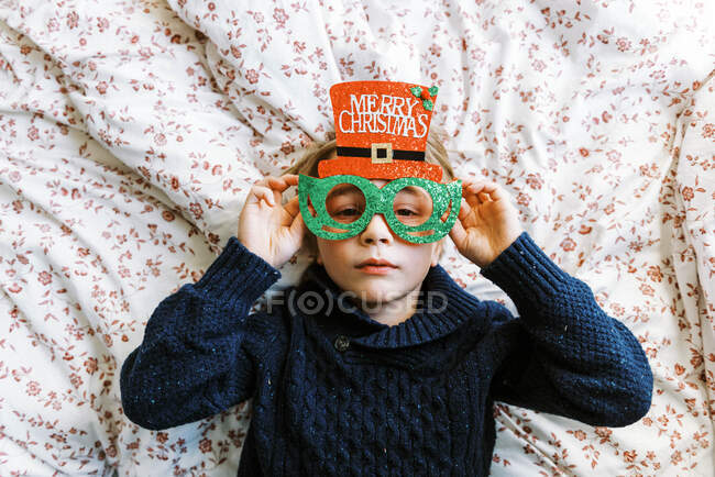 Little boy in a knit sweater with Christmas glasses lying on bed — Stock Photo