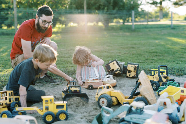 Um pai e seus filhos brincando em uma caixa de areia com caminhões juntos — Fotografia de Stock