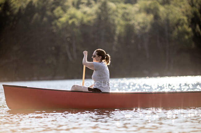 Woman paddles red canoe across lake in sunshine in Maine woods — Stock Photo