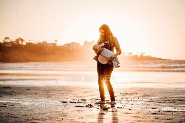 Mãe segurando bebê amamentando na praia durante o pôr do sol no outono — Fotografia de Stock