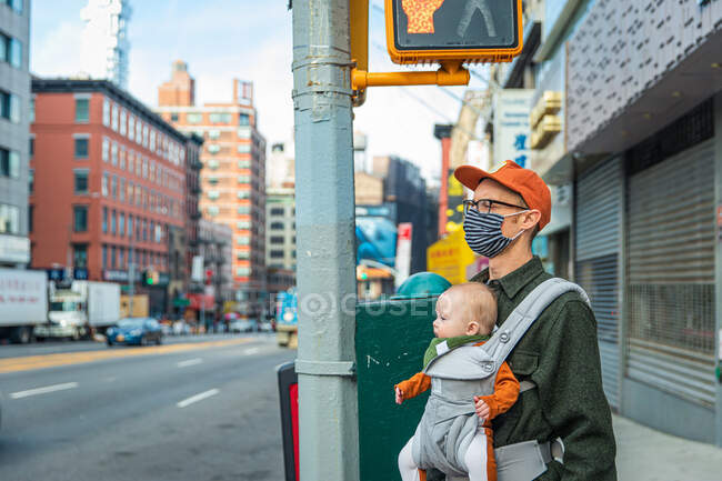 Padre con mascarilla que lleva a una niña mientras está de pie en la acera de la ciudad durante el brote de COVID-19 - foto de stock