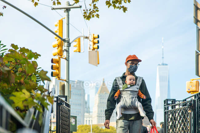Padre llevando linda hija en portabebés mientras camina en la ciudad durante COVID-19 - foto de stock