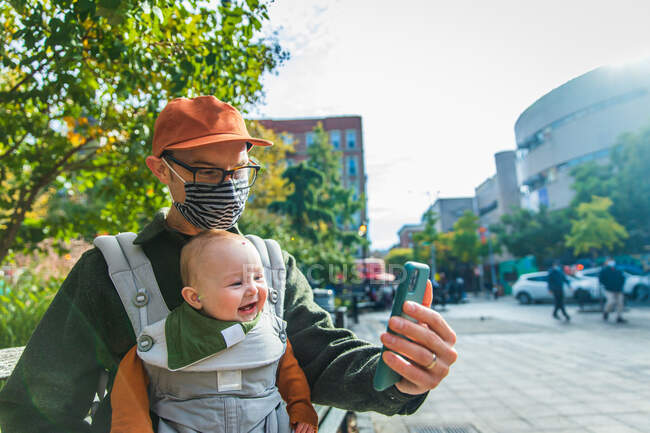 Man with happy baby daughter video calling through smart phone while sitting on bench in city during COVID-19 — Stock Photo