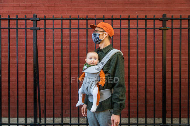 Father wearing face mask carrying cute daughter in baby carrier while standing by fence during coronavirus crisis — Stock Photo