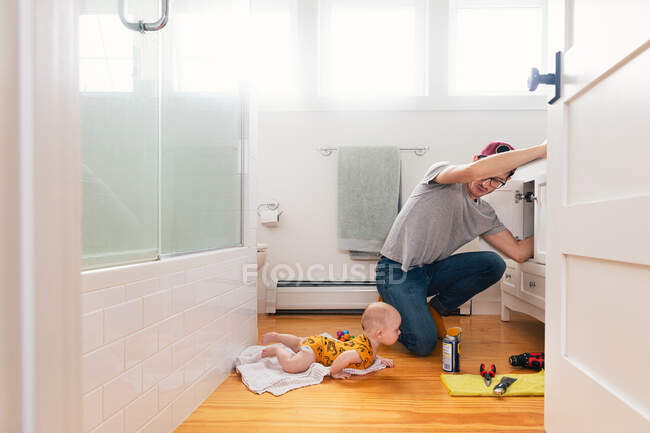 Man fixing sink while daughter lying on hardwood floor in kitchen — Stock Photo