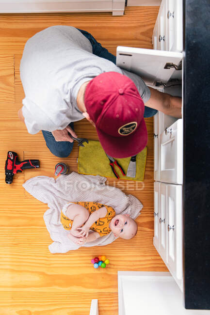 Vista aérea del padre que fija el fregadero de la cocina mientras que la muchacha del bebé que miente en piso de madera dura en el país - foto de stock