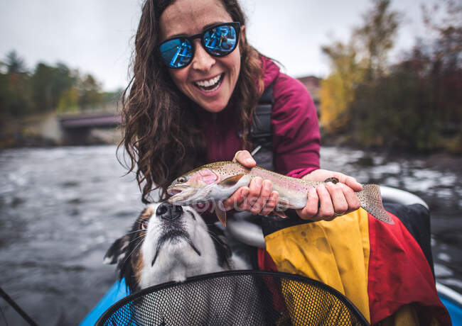 Mulher segurando uma truta arco-íris em barco com cão farejando — Fotografia de Stock