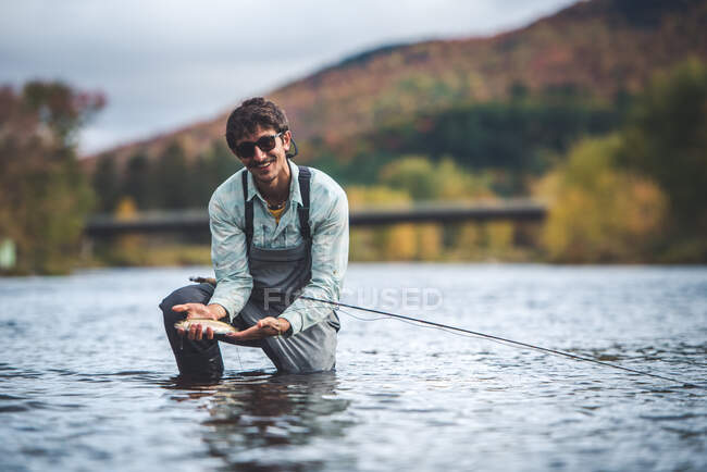 L'homme tient la truite arc-en-ciel dans une rivière avec du feuillage en arrière-plan — Photo de stock