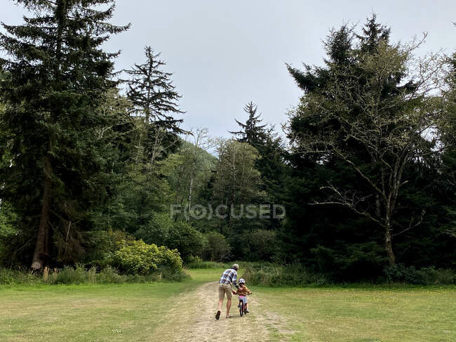 Un padre enseña a su hija a montar en bicicleta en un campo de hierba. - foto de stock