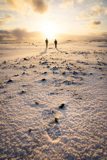 Dos personas caminando al amanecer en la playa de arena negra en Islandia en invierno - foto de stock