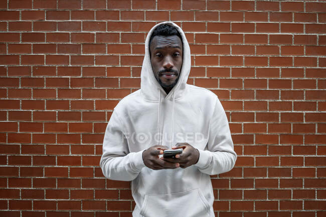 Foto frontal del hombre africano con gesto serio y capucha usando el teléfono inteligente en la ciudad - foto de stock
