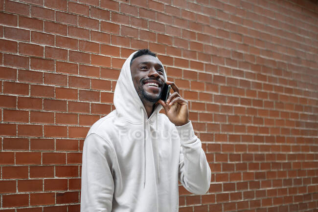 Seitenaufnahme eines Afrikaners mit glücklicher Geste und Kapuze per Smartphone in der Stadt — Stockfoto