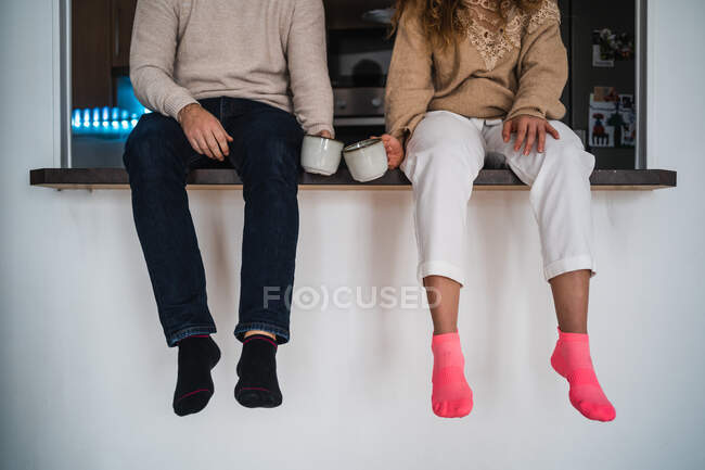 Anonymous couple with cups of hot drink sitting on table in kitchen — Fotografia de Stock