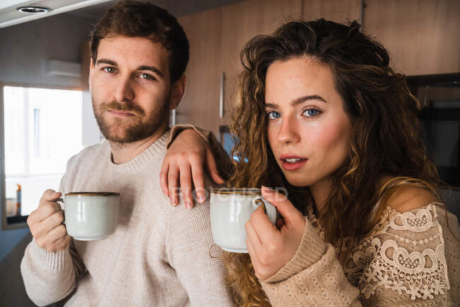 Молода пара з гарячим напоєм на кухні вдома дивиться на камеру — стокове фото