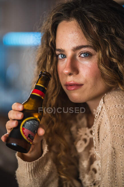 Mujer joven bebiendo cerveza en casa y mirando a la cámara - foto de stock