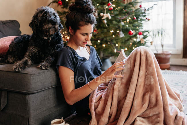 Smiling young woman reading with her dog in front of Christmas tree — Stock Photo