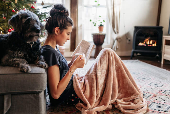 Young woman reading by the fireplace with dog, drinking a hot beverage — Stock Photo
